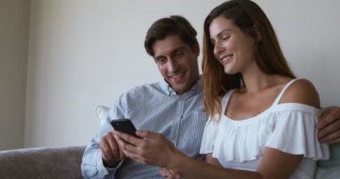 Kafkasyalı bir çiftin birlikte bir dairede otururken, bir koltukta otururken, akıllı bir telefon tutarken, üzerinde ağır çekimde bir şeyler izlerken yan görüntüsü.