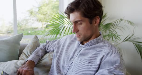一个白人男人坐在公寓里 坐在沙发上 拿着智能手机 看着相机 慢吞吞地微笑 侧视近景 — 图库视频影像