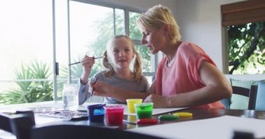 Mutlu, beyaz bir kadın kızıyla birlikte evde vakit geçiriyor, oturma odasında bir masada oturuyor, resim yapıyor, gülümsüyor ve birbirlerine bakıyor, burnunu boyuyor, sosyal mesafe koyuyor ve karantinada kendini izole ediyor.