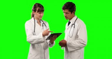 Önde iki beyaz erkek ve kadın doktorun laboratuvar önlüğü ve stetoskop giyip yeşil ekran arka planda tartışıp tablet kullanırken görüntüsü var..