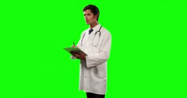 Önde, laboratuvar önlüğü ve steteskop giyen beyaz bir erkek doktorun elinde bir dosya ve yeşil ekran arka plan üzerine notlar tutarken görüntüsü var..