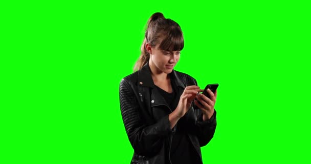 一个有着一头乌黑长发 一件皮夹克 面带微笑并在绿色屏幕背景上使用智能手机的快乐的高加索女人的前视特写 — 图库视频影像