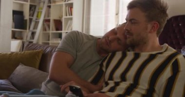 Kafkasyalı mutlu bir erkeğin, aynı cinsiyetten bir çiftin oturma odasında, kanepede oturup, kucaklaşıp televizyon izlemesini, uzaktan kumandayı elinde tutmasını.