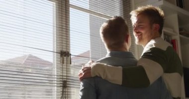 Kafkasyalı mutlu bir erkek, aynı cinsiyetten bir çiftin oturma odalarında dinlenip, güneşin altında birbirlerine sarılmalarını, gülümsemelerini ve pencereden dışarı bakmalarını izliyorlar. Karantina karantinasında sosyal mesafeler ve kişisel izolasyon yavaş çekimde