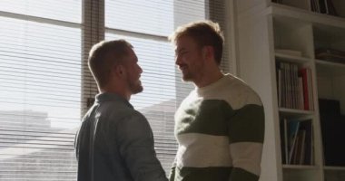 Mutlu beyaz erkek aynı cinsiyetten çift oturma odalarında güneşleniyorlar, pencerenin kenarında duruyorlar birbirlerine gülümsüyorlar ve öpüşüyorlar. Karantina karantinasında sosyal mesafeler ve kişisel izolasyon yavaş çekimde