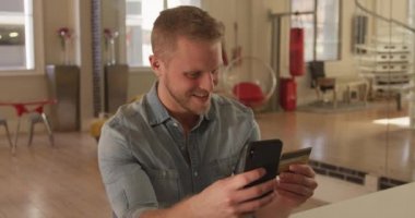 Kafkasyalı bir adam evinde oturmuş, akıllı telefonunu kullanıyor ve elinde kredi kartıyla gülümsüyor. Karantina karantinasında sosyal mesafeler ve kişisel izolasyon yavaş çekimde