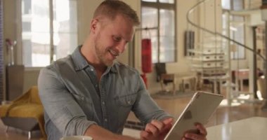 Beyaz adam evinde, oturma odasında tablet bilgisayar kullanarak çalışıyor ve gülümsüyor. Karantina karantinasında sosyal mesafeler ve kişisel izolasyon yavaş çekimde