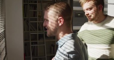 Yandan bakan mutlu beyaz erkek aynı cinsiyetten çift oturma odalarında dinleniyor, güneşin altında duruyor, birbirlerine gülümsüyor ve pencereden dışarı bakıyorlar.