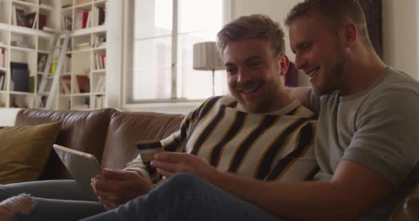 二人はソファに座ってオンラインで支払いをします一方はタブレットコンピュータを持っていますもう一方はクレジットカードを持っていて両方とも笑顔です — ストック動画