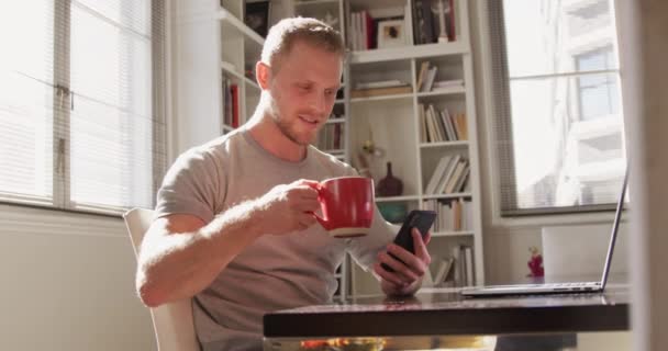 Nějaký běloch doma ve svém obýváku, sedí za stolem na slunci, používá smartphone a popíjí šálek kávy, na stole před sebou má přenosný počítač. Sociální distancování a izolaci v karanténě, ve zpomaleném filmu
