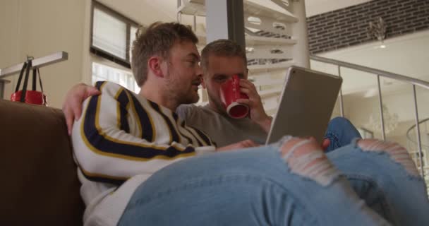 幸せな白人男性の低い角度ビュー同じセックスカップルリラックスして自宅で彼らのリビングルームで ソファに座って 話して タブレットコンピュータを一緒に見て 一人の男飲むコーヒーカップ — ストック動画
