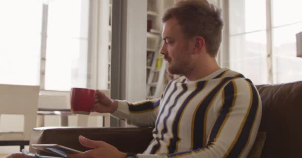 リビングルームでリラックスしている白人男性の近くにあり タブレットコンピュータを使用してソファに座ってコーヒーを飲んでいます 社会的距離と隔離された封鎖の自己分離スローモーションで — ストック動画