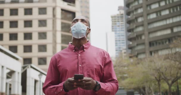 アフリカ系アメリカ人の男性は 大気汚染や海綿状ウイルスに対して顔マスクを着用し 日中街を歩き回っています19コロナウイルスは 彼のスマートフォンを使用して街を探索します — ストック動画