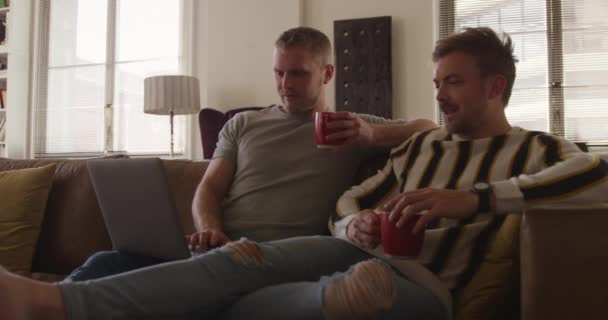 幸せな白人男性同じセックスカップル自宅で一緒にリラックスしたリビングルームで ソファに座って 話して コーヒーを飲んでいます 社会的距離と隔離された封鎖の自己分離スローモーションで — ストック動画