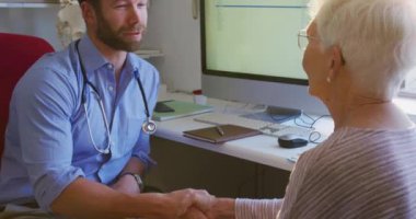 Beyaz erkek doktor mavi gömlek ve steteskop giyiyor, bir huzurevinde ameliyatta çalışıyor, Coronavirus covid19 salgını sırasında önünde oturan yaşlı beyaz bir kadını dinliyor..