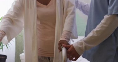 Mavi önlük giyen Kafkasyalı bir hemşire, yaşlı bir Afrikalı Amerikalı kadına Coronavirus Covid19 salgını sırasında bir huzurevinde yürüyen bir çerçeveyle yürümesinde yardım ediyor..