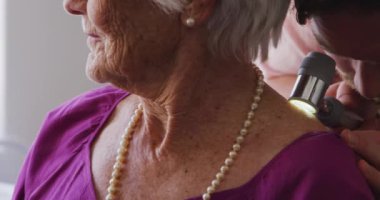 Pembe gömlek ve steteskop giyen beyaz bir erkek doktoru yakın plan, bir huzurevinde ameliyatta çalışıyor, Coronavirus covid19 salgını sırasında önünde oturan yaşlı beyaz bir kadını muayene ediyor..