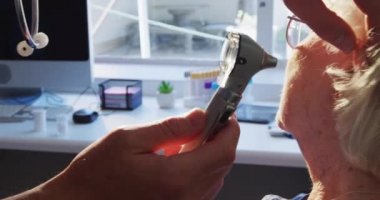 Pembe gömlek ve steteskop giyen beyaz bir erkek doktoru yakın plan, bir huzurevinde ameliyatta, Coronavirus covid19 salgını sırasında, yaşlı bir beyaz kadın hastanın kulağını muayene ediyor..