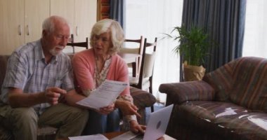 Üst düzey Kafkasyalı çift evde birlikte vakit geçiriyorlar, kanepede oturup idari kağıtları okuyor ve açıklıyorlar ve Coronavirus covid19 salgını sırasında akıllı telefonlarını kullanıyorlar..