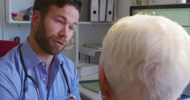 Beyaz erkek doktor mavi gömlek ve steteskop giyiyor, bir huzurevinde ameliyatta çalışıyor, önünde oturan yaşlı beyaz bir kadınla görüşüyor, Coronavirus covid19 salgını sırasında tedavisini anlatıyor..
