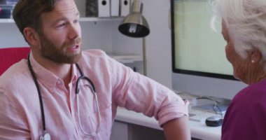 Beyaz erkek doktor pembe gömlek ve steteskop giyiyor, bir huzurevinde ameliyatta çalışıyor, Coronavirus covid19 salgını sırasında önünde oturan yaşlı beyaz bir kadınla konuşuyor..