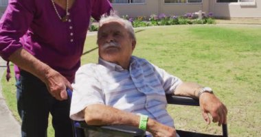Üst düzey beyaz kadın güneşli bir günde, tekerlekli sandalyede, bir huzurevinin parkında oturan kıdemli beyaz bir adamla yürüyor..