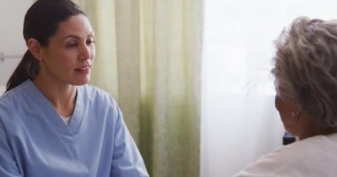 Mavi önlük giyen beyaz bir hemşire, yaşlı bir Afrikalı Amerikalı kadınla konuşuyor, tekerlekli sandalyede oturuyor, huzurevinde yaşıyor, Coronavirus covid19 salgını sırasında ona destek oluyor..