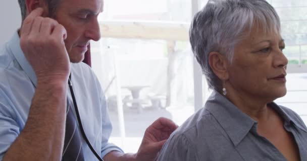 老人ホームで手術に従事している白人男性医師は 聴診器を使用して高齢の混合人種の女性患者を調べ コロナウイルスの感染症19パンデミックの間 — ストック動画