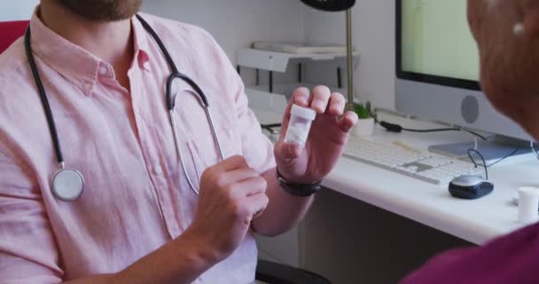 ピンクのシャツと聴診器を身に着けている白人男性医師 老人ホームで彼の手術で作業 彼の前に座っているシニア白人女性への治療方法を説明 コロナウイルスの流行の間に19 — ストック動画