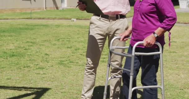 在一家养老院工作的白种人医生 与一名头脑清醒的老年白种人女病人在一个阳光灿烂的日子里 在一场大流行病中散步 展示了她的检查结果 — 图库视频影像