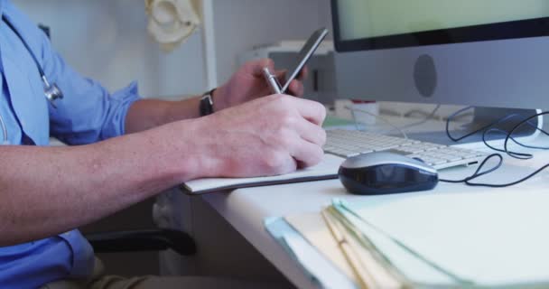 白人男医生穿着蓝色衬衫和听诊器 在一家养老院做手术 用智能手机和写便条 — 图库视频影像