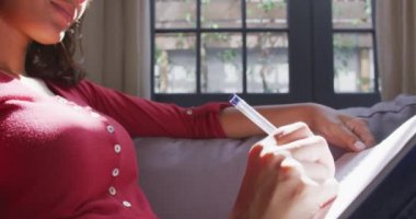 Karışık ırk kadını evinde keyif alıyor, kanepede uzanıyor, kulaklıkla müzik dinliyor, yazıyor ve gülümsüyor, sosyal uzaklık ve karantina hücresinde kendini izole ediyor, yavaş çekimde.