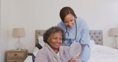 Karışık ırktan bir kadın tekerlekli sandalyede oturan melez bir hemşire tarafından ziyaret ediliyor, aynaya bakıyor, sosyal uzaklık ve karantina tecridinde, yavaş çekimde