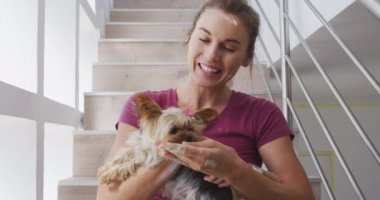 Beyaz bir kadın, Coronavirus covid 19 salgını sırasında karantinada evini, sosyal mesafeyi ve kendini izolasyonunu yenileyerek evde vakit geçiriyor. Köpeğini tedavi ediyor, merdivenlerde oturuyor ve ağır çekimde gülümsüyor.