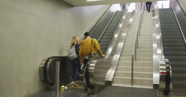 Sıradan kıyafetler giyen, metro istasyonundan ayrılan, yürüyen merdivende durup gülümseyen ve konuşan beyaz bir çiftin arka görüntüsü..