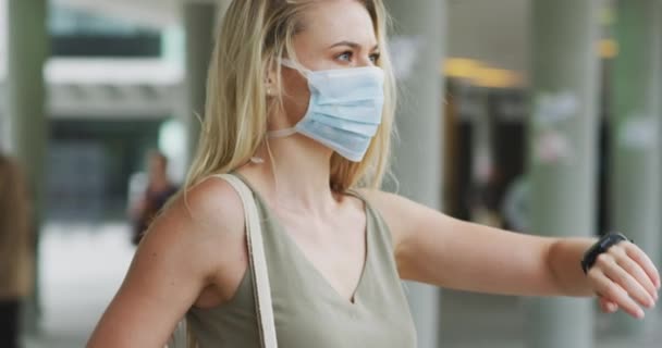 コロナウイルスに対して顔マスクを身に着けている白人女性は スマートウォッチを使用して 日中に街の通りで19をCovidし 男性が背景に通過している間 — ストック動画