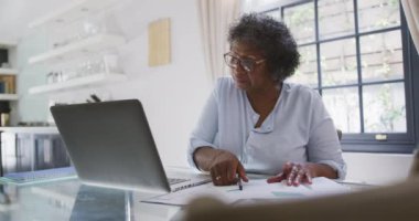 Üst düzey melez kadın evinde, masada oturup dizüstü bilgisayar kullanıyor, bir parça kağıda yazıyor, sosyal uzaklık ve kendini karantinaya almış, ağır çekimde.