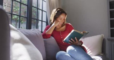 Karışık ırktan olan bir kadın, kanepede oturup kitap okuyarak eğleniyor ve gülümsüyor, sosyal uzaklaşıyor ve kendini karantinaya alıyor, ağır çekimde.