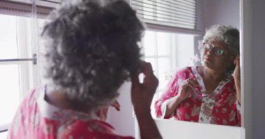 Kıdemli bir Afrikalı Amerikalı kadın Coronavirus covid 19 salgını sırasında karantinada karantina ve kişisel izolasyon ile evde vakit geçiriyor. Aynaya bakıyor ve yavaşça yüzüne dokunuyor.