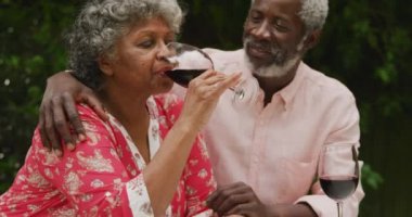Kıdemli bir Afrikalı Amerikalı çift, Coronavirus covid 19 salgını sırasında karantinada, sosyal mesafeyi ve kendilerini izole ederek birlikte bahçelerinde vakit geçiriyorlar, ellerinde kırmızı şarap kadehleri ve yavaş çekimde içiyorlar.