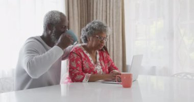 Üst düzey bir Afrikalı Amerikalı çift, Coronavirus covid 19 salgını sırasında karantinada birlikte vakit geçiriyorlar. Masada oturuyorlar. Dizüstü bilgisayar kullanan kadın, elinde bardak tutan ve içen adam.