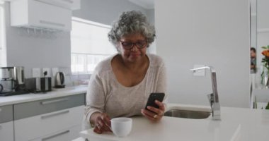 Üst düzey bir Afrikalı Amerikalı kadın, Coronavirus covid 19 salgını sırasında karantina altında, evde birlikte vakit geçiriyor. Akıllı telefon kullanıyor, elinde bardak tutuyor ve yavaş çekimde içiyor.