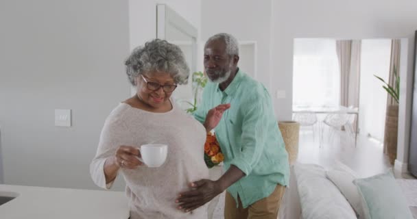 コロナウイルスの流行の間に隔離されたロックダウンで社会的距離と自己隔離を一緒に家で過ごす上級アフリカ系アメリカ人のカップル 遅い動きで女性に花を与える男 — ストック動画