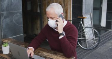 Orta yaşlı beyaz bir adam kahve terasında bir masada oturuyor, Coronavirus 'a karşı maske takıyor, covid 19, akıllı telefon ve dizüstü bilgisayar kullanıyor, ağır çekimde..