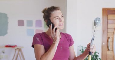 Beyaz bir kadın evinde vakit geçiriyor, evini yeniliyor, sosyal uzaklık ve karantinada kendini izole ediyor, Coronavirus covid 19 salgını sırasında, elinde boya silindiri tutarak, akıllı bir telefonla konuşuyor.