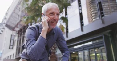 Orta yaşlı, beyaz bir adam gün boyunca şehir sokaklarında dolaşıyor gri bir gömlek giyiyor, akıllı telefonuyla gülümsüyor ve ağır çekimde konuşuyor..