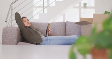Beyaz bir kadın, Coronavirus covid 19 salgını sırasında karantinada, evinde vakit geçirir. Kanepede oturur, elinde şişe tutar ve ağır çekimde içerdi.