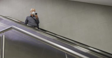 Coronavirus 'a karşı yüz maskesi takan orta yaşlı bir adam, Covid 19, metro istasyonunda yürüyen merdiven kullanıyor ve akıllı telefonunu yavaş çekimde kullanıyor..