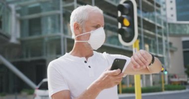 Orta yaşlı beyaz bir adam gün boyunca şehir sokaklarında dolaşır, Coronavirus 'a karşı yüz maskesi takar, covid 19, akıllı saatini ve akıllı telefonunu yavaş çekimde kullanır..