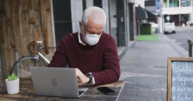 Orta yaşlı beyaz bir adam kahve terasında bir masada oturuyor, Coronavirus 'a karşı maske takıyor, covid 19, akıllı telefon ve dizüstü bilgisayar kullanıyor, ağır çekimde..